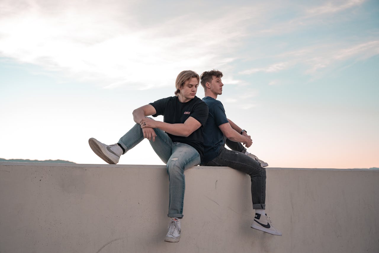 Two young men sitting back to back on a wall under a clear evening sky.