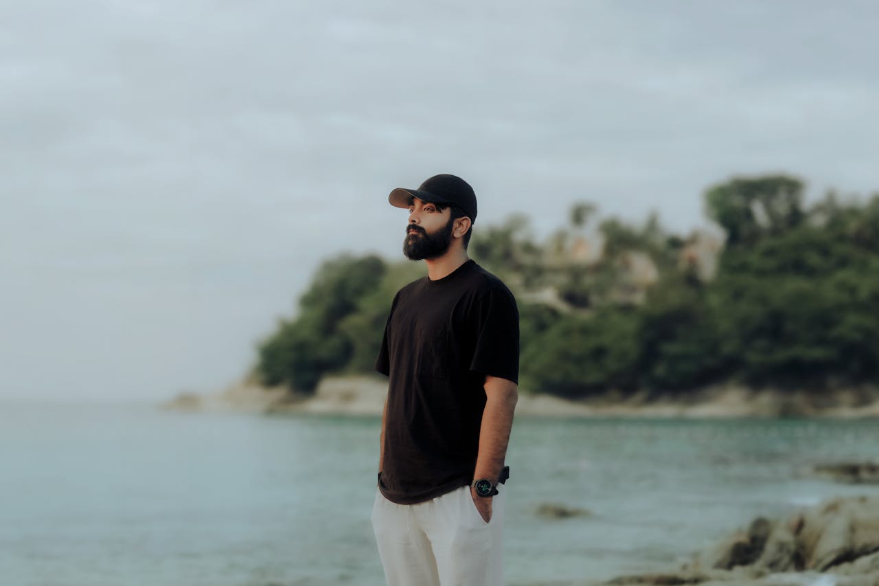 A man in casual attire stands thoughtfully by a serene beach, capturing a moment of reflection.